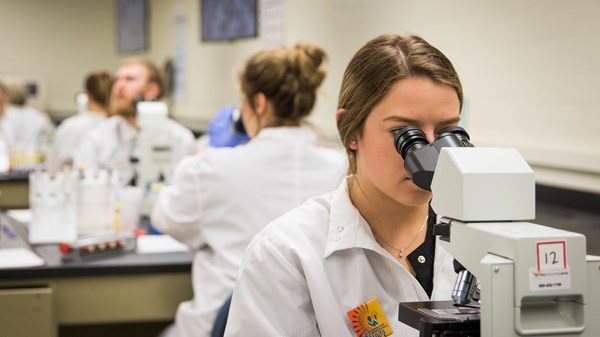 A photo of a female student in a white lab coat looking through a microscope.