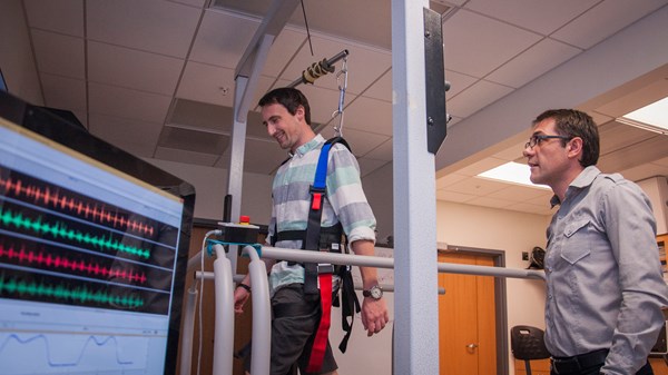 A photo of a male researcher observing a male subject walking on a machine in a laboratory.