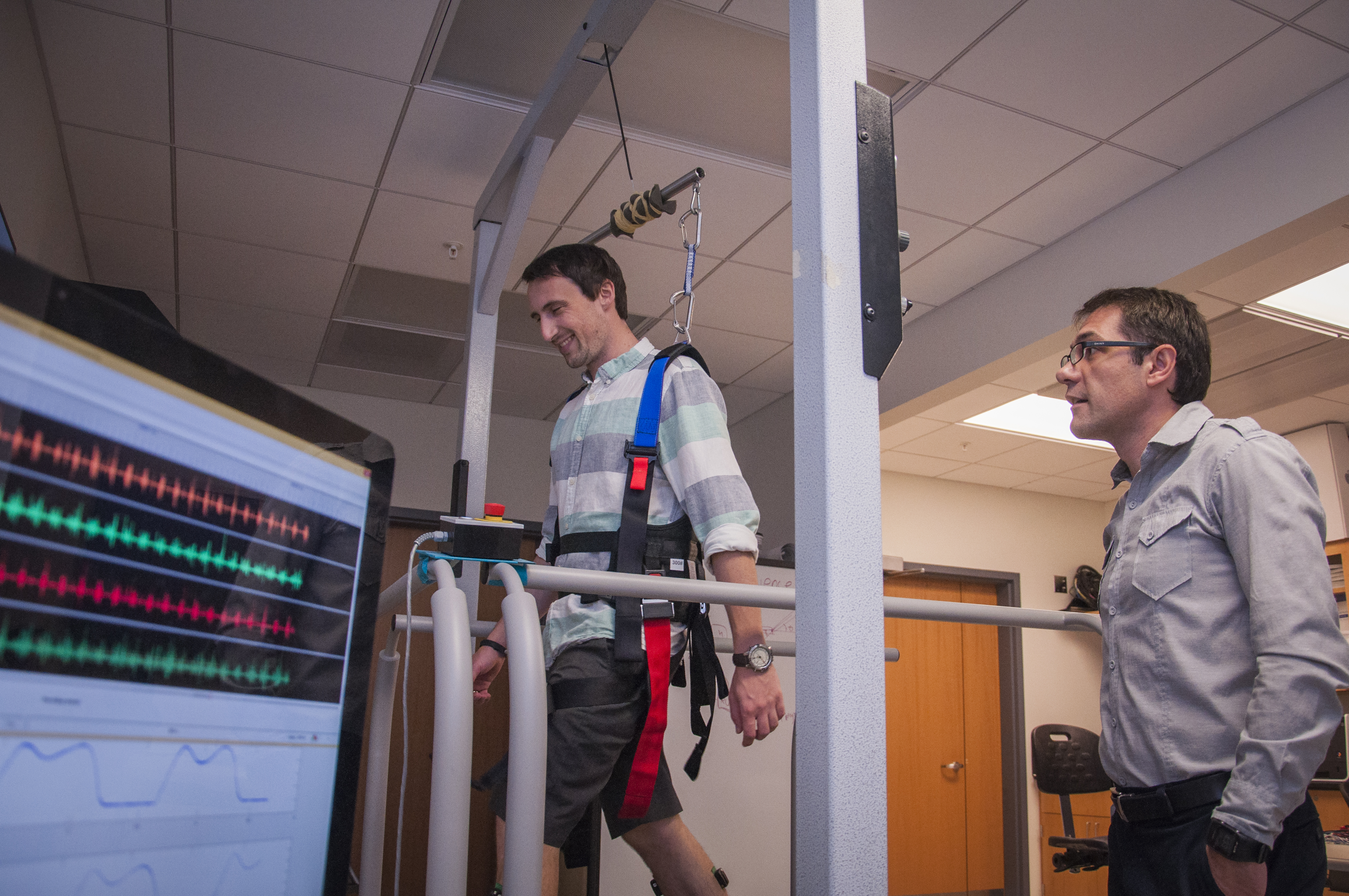 A photo of a male researcher observing a male subject walking on a machine in a laboratory.