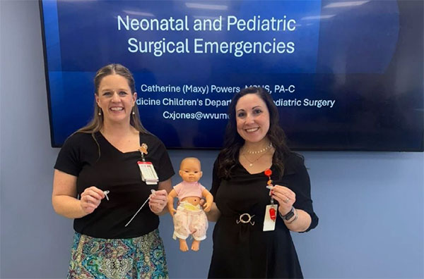 Two Physician Assistant female preceptors  holding a baby doll in front of a screen that reads, "Neonatal and Pediatric Surgical Emergencies"