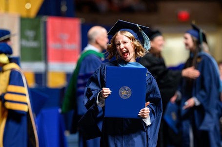 WVU MOT graduate, Ava, walking across the stage at her graduation ceremony with a smile showing off her degree.
