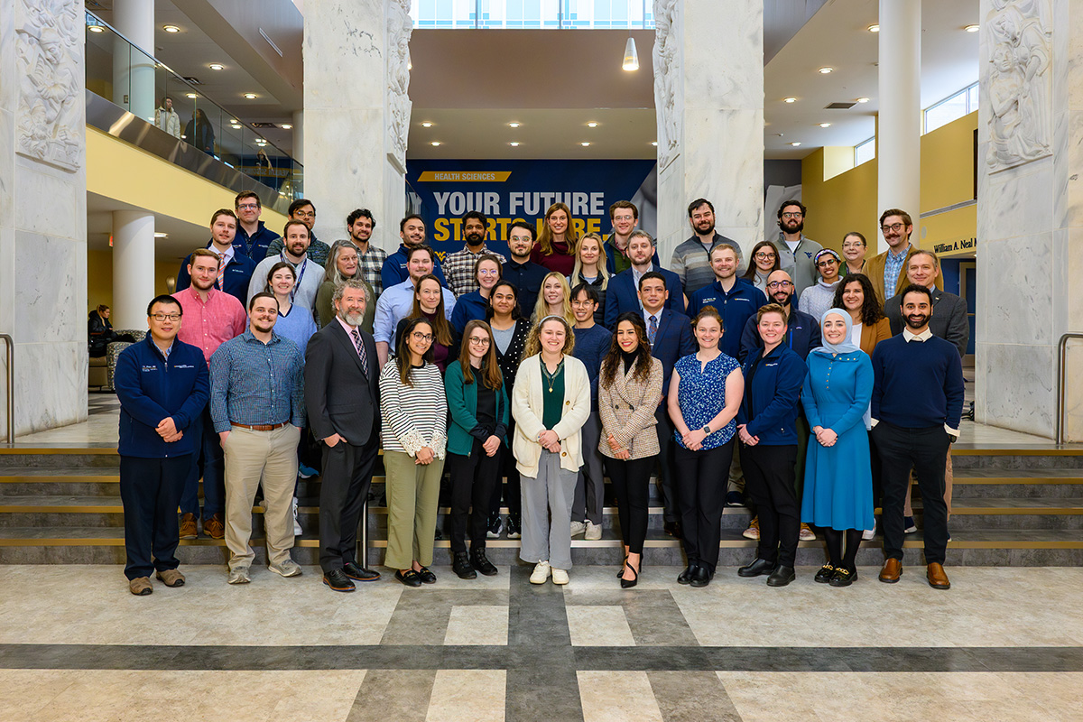 A large group of diverse medical professionals posing for a group portrait in a bright, modern atrium. They are standing on a wide set of steps in front of two large marble pillars. In the background, a blue wall features the West Virginia University logo and the text "Your future starts here."